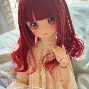 Puede incluir: Una muñeca con el pelo rojo y los ojos rojos que lleva un vestido blanco con encaje y ribetes dorados. La muñeca lleva un collar de perlas alrededor del cuello y el texto "Strawberry Rin" está escrito en el vestido.