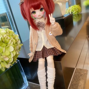 May include: A red-haired doll wearing a brown cardigan, a white shirt, a plaid skirt, and white knee-high socks. The doll is holding a peace sign with her right hand.