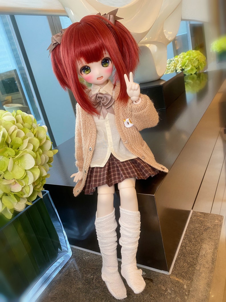 May include: A red-haired doll wearing a brown cardigan, a white shirt, a plaid skirt, and white knee-high socks. The doll is holding a peace sign with her right hand.