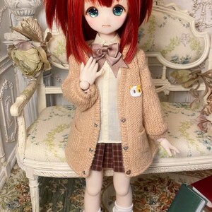 May include: A doll with red hair in pigtails wearing a brown cardigan, a white shirt with a brown bow tie, a plaid skirt, and white knee-high socks.