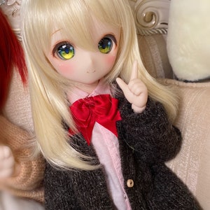 May include: A doll with long blonde hair wearing a brown cardigan, a pink shirt with a red bow tie, and a book in her hand.