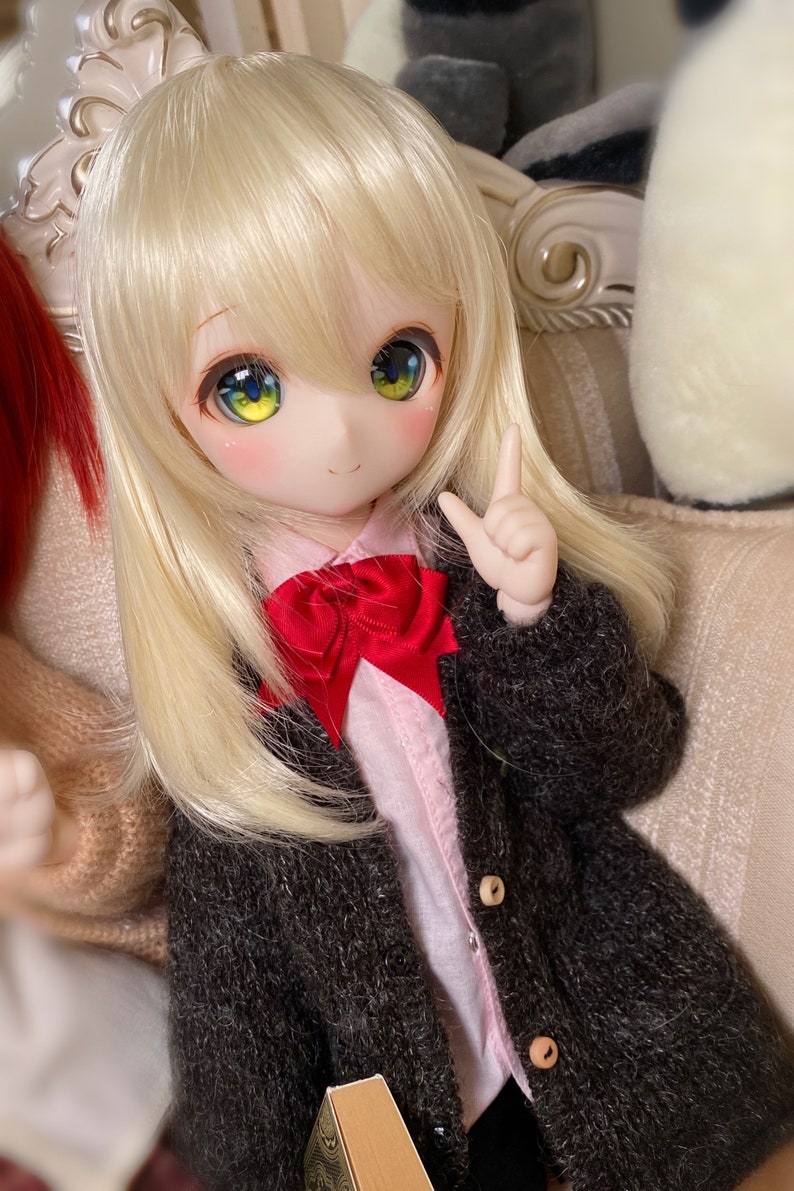 May include: A doll with long blonde hair wearing a brown cardigan, a pink shirt with a red bow tie, and a book in her hand.