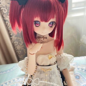 Op de afbeelding: Een pop met rood haar en zwarte strikken in haar haar draagt een witte jurk met een zwart-wit gepatenteerde rok. De pop houdt een bruine en gouden strik in haar hand. De pop zit op een gepatenteerd oppervlak met een parelketting om haar nek.