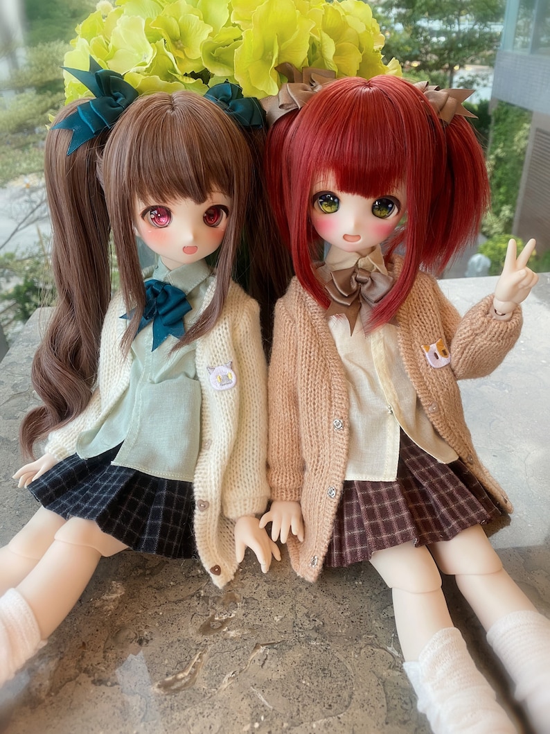 May include: Two dolls wearing school uniforms with brown and red hair, brown and white cardigans, and plaid skirts. The doll on the left has a teal bow and the doll on the right has a brown bow.