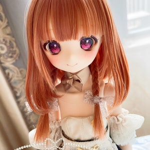 May include: A doll with long, straight, auburn hair styled in two braids. The doll has large, brown eyes with pink accents and is wearing a white lace dress with a brown bow.