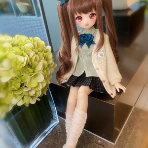 May include: A doll with long brown hair styled in pigtails with blue bows. She is wearing a white cardigan, a green shirt, a black and white plaid skirt, and white knee-high socks. The doll is sitting on a black surface.