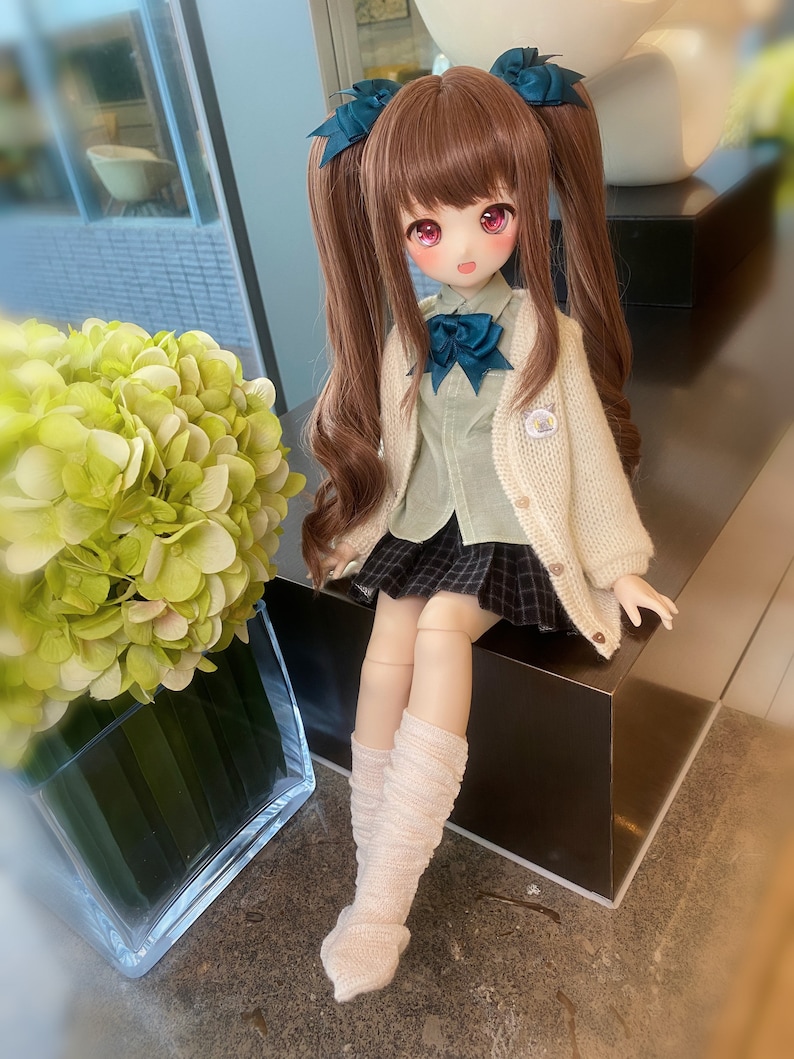 May include: A doll with long brown hair styled in pigtails with blue bows. She is wearing a white cardigan, a green shirt, a black and white plaid skirt, and white knee-high socks. The doll is sitting on a black surface.