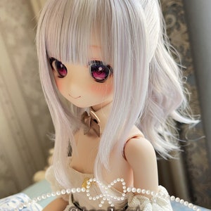 May include: A doll with long, white hair styled in a ponytail with a white bow. The doll is wearing a white dress with a brown bow around the neck and a pearl necklace. The doll's eyes are pink and she has a light pink blush on her cheeks. The text "Stardust Patricia (short)" is written below the doll.