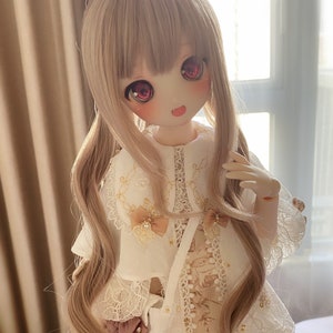 Puede incluir: Una muñeca con el pelo largo, ondulado y castaño claro, que lleva un vestido blanco con ribetes de encaje y un lazo rosa en el pelo. La muñeca tiene ojos grandes y rojos y una expresión dulce.