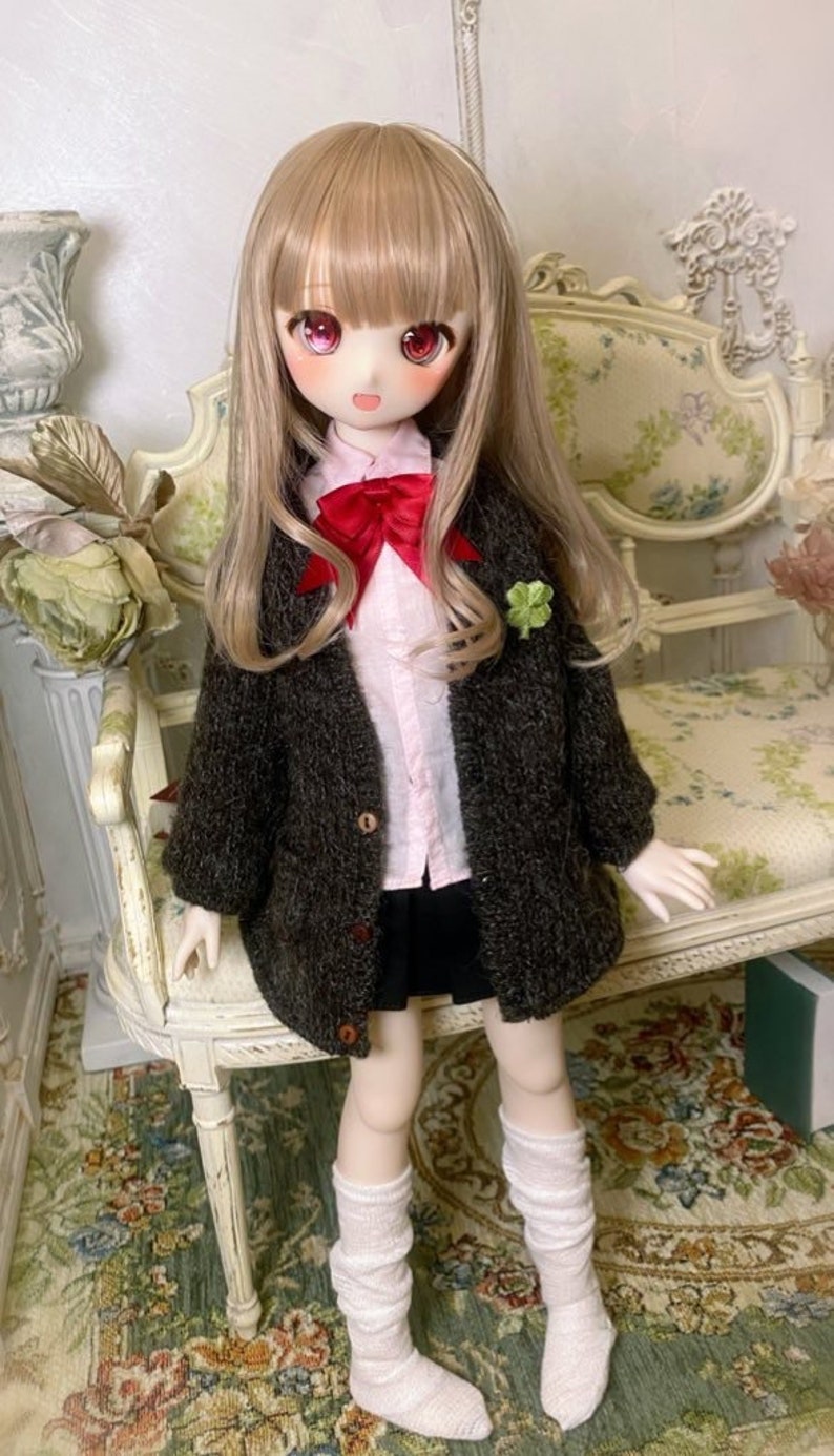 May include: A doll with long blonde hair wearing a brown cardigan, a pink shirt, a black skirt, and white knee-high socks. The doll is sitting on a white chair with a floral pattern.