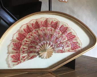 May include: A framed, red and gold lace fan with a wooden handle. The fan is displayed on a wooden surface.