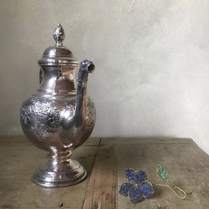 Op de afbeelding: Een zilverkleurige theepot met bloemmotieven, een gebogen tuit en een decoratief deksel. Daarnaast een blauw-gouden bloemvormige broche met een groen blad. De objecten staan op een houten oppervlak.