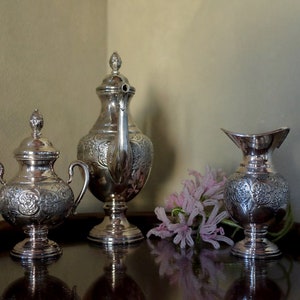 May include: A silver tea set with intricate floral designs on a wooden tray. The set includes a teapot, creamer, and sugar bowl.