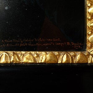 May include: A close-up of a black wooden frame with gold accents. The frame has a gold inscription that reads "Sones Himaly Olelsige bielone been lens, Asantot festett k&eacute;pestre, maroli Gempeh B. Drexia- 105- "