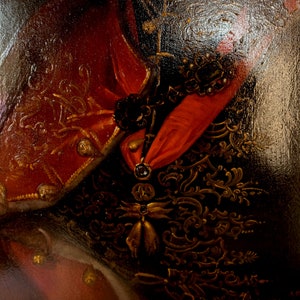 May include: Close-up of a red and gold embroidered fabric with a black and gold patterned background. The fabric has a textured surface and is likely part of a historical costume.
