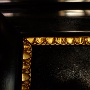May include: Close-up of a black picture frame with a gold ornate border. The frame is made of wood and has a smooth, polished finish.