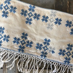 May include: A white table runner with a blue and white embroidered pattern of snowflakes. The runner has a white fringe on the bottom edge.