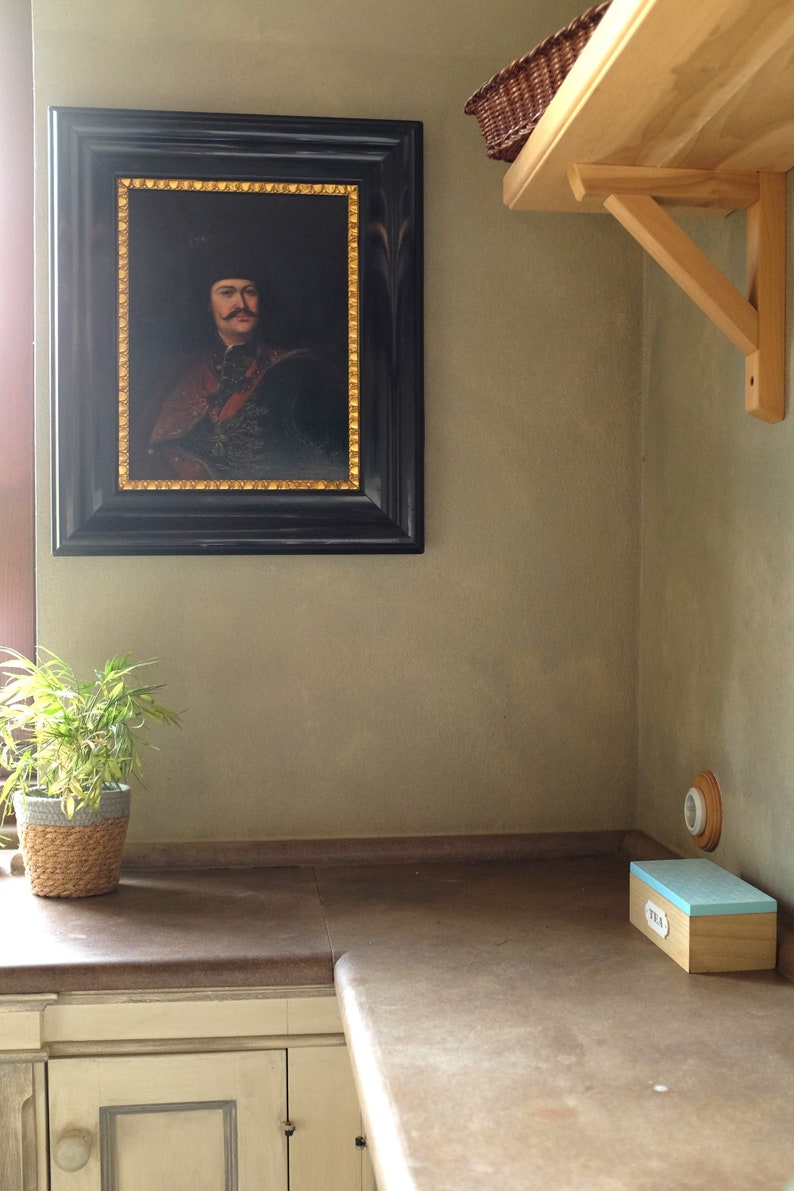 May include: A framed portrait of a man in a black hat and a dark red and gold jacket. The portrait is hanging on a light green wall. A wooden shelf with a wicker basket is above the portrait. A small potted plant is on a brown countertop below the portrait.