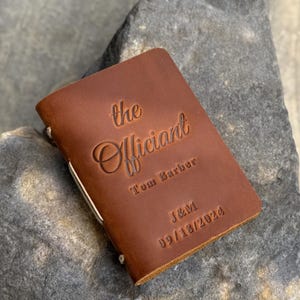 May include: A brown leather notebook with the embossed text "the Officiant" and the name "Tom Barber". The notebook also features the initials "J&M" and the date "09/18/2024". A cream-colored string binds the pages.
