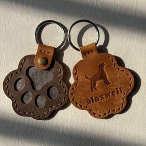 May include: Two paw-shaped leather keychains with metal rings. The left keychain is dark brown with a cutout paw print. The right keychain is tan leather, embossed with a dog silhouette and the name Maxwell, featuring a brass-colored rivet.