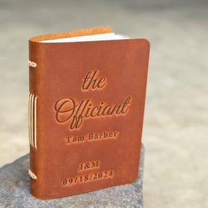 May include: A small, brown leather-bound book with the words "the Officiant" embossed on the cover. The name "Tom Barber" and the date "09/18/2024" are also visible. The book is held together with white thread.