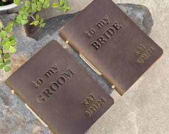 May include: Two brown leather journals with the text "to my groom" and "to my bride" engraved on the front. The journals also have the initials "K&J" and the date "9/21/24" engraved on the front.