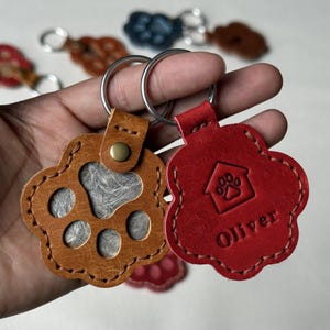 Puede incluir: Dos llaveros de cuero con forma de pata y anillas metálicas. El llavero color canela tiene un diseño de pata recortada con pelo gris visible en el interior. El llavero rojo está grabado con un icono de casa, una pata y el nombre Oliver.