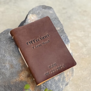 May include: A brown leather notebook with the word "Officiant" and the name "Liam Seal" embossed on the cover. The date "08/22/2024" is also visible. The notebook is resting on a gray stone.