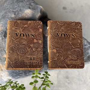 Könnte beinhalten: Zwei braune Leder-Gelübde-Bücher mit floraler Prägung. Das Wort "VOWS" ist auf der Vorderseite jedes Buches geprägt. Die Initialen "F&E" und das Datum "09/06/24" sind ebenfalls auf der Vorderseite jedes Buches geprägt. Die Bücher sind mit einer weißen Schnur gebunden.