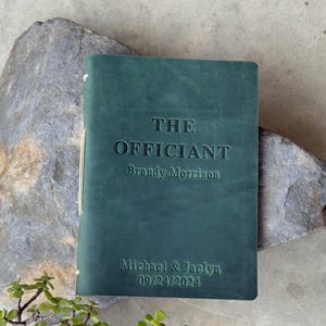 May include: A dark teal leather-bound book with the embossed text "THE OFFICIANT" and the name "Brandy Morrison". Below, the names "Michael & Jaclyn" and the date "09/24/2024" are also embossed. The book is bound with a cream-colored string.