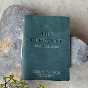 Puede incluir: Un libro encuadernado en cuero verde oscuro con el título "THE OFFICIANT" y el nombre "Brandy Morrison" en relieve en la portada. El texto "Michael & Jaclyn 09/24/2024" también está en relieve en la portada.