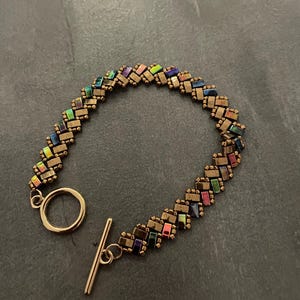 Peut inclure: Un bracelet de perles avec un fermoir à bascule. Le bracelet présente de petites perles rectangulaires aux couleurs irisées variées, dont le vert, le violet et le rose, serties dans un métal de couleur bronze. Le fermoir est doré.