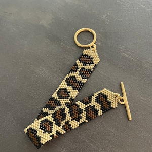 Puede incluir: Un brazalete de cuentas con un diseño de estampado de leopardo. El brazalete tiene un cierre dorado y un cierre de palanca dorado.
