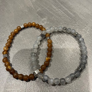 Peut inclure: Deux bracelets en perles, l'un avec des perles brunes et dorées et l'autre avec des perles grises. Les deux bracelets ont des perles carrées argentées.