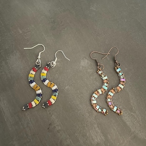 Peut inclure: Deux paires de boucles d'oreilles faites à la main. Une paire présente un motif en zigzag coloré avec des perles rouges, blanches, jaunes, bleues et noires. L'autre paire a un motif en zigzag avec des perles irisées et marron. Les deux paires ont des crochets d'oreilles argentés et cuivrés.