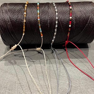 May include: Four beaded bracelets with different colored beads on a spool of black thread. The bracelets are tied with white, gray, and red cords.