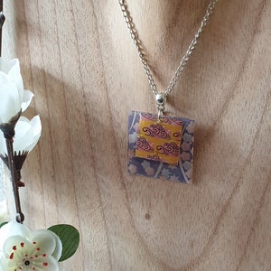 Può includere: Una collana con catena d'argento con un ciondolo quadrato che presenta un design a strati di carta fantasia gialla e viola. Il ciondolo ha un motivo a nuvole rosa e gialle sullo strato superiore e un motivo floreale viola e bianco sullo strato inferiore.