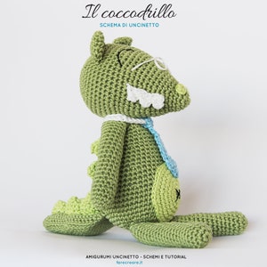 May include: A green crocheted alligator wearing glasses and a blue tie. The alligator is sitting on a white background. The text "Il coccodrillo" and "Schema di uncinetto" is above the alligator. The text "AMIGURUMI UNCINETTO - SCHEMI E TUTORIAL" and "farecreare.it" is below the alligator.