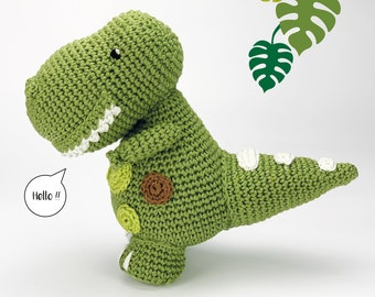 Op de afbeelding: Groen gehaakt dinosaurus speelgoed met witte stippen en bruine accenten. De dinosaurus staat op een wit oppervlak en heeft een spraakballon met de tekst "Hello!!".