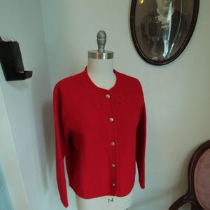 May include: A bright red long-sleeve cardigan sweater featuring a round neckline and five gold-toned metal buttons down the front. The knit garment has a subtle textured pattern around the collar area and is displayed on a mannequin torso.