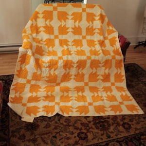 May include: A rectangular quilt featuring a repeating geometric pattern in bright orange and off-white fabric. The design consists of interconnected squares and triangular shapes arranged in a grid, with visible stitching throughout the surface.