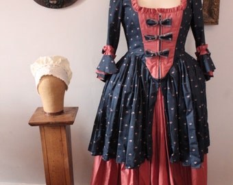 May include: A historical costume dress on a mannequin. The dress features a navy blue bodice with small white polka dots and a red panel with black bows. The skirt is red with a ruffled hem. A white cap sits on a wooden stand.