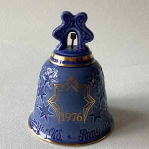 May include: A blue ceramic bell with gold detailing and star patterns. The bell features the year "1976" in gold and the words "Ringing in the Old" in blue. The bell has a decorative handle.
