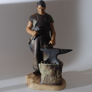 May include: A ceramic figurine of a blacksmith, wearing a dark apron and holding a hammer. The blacksmith stands next to a black anvil on a stone base. The figurine is painted in shades of brown and tan.