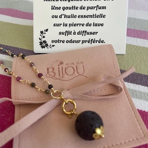 Pode incluir: Um colar dourado com um pingente de pedra de lava cinza escuro e contas coloridas. O colar é exibido com uma bolsa rosa e um cartão branco com texto em francês que diz "Alliez élégance et bien-être".