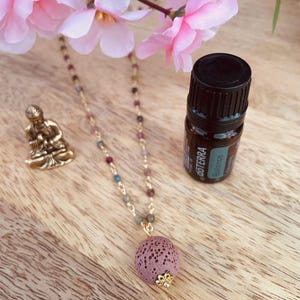 Può includere: Una collana con catena dorata con un ciondolo in pietra lavica viola e perline colorate. Una piccola statua di Buddha dorata e una bottiglia di olio essenziale sono anche sulla superficie di legno. Fiori rosa sullo sfondo.