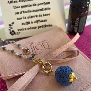Può includere: Una collana color oro con un ciondolo in pietra lavica blu e piccole perline blu. La collana è esposta con una piccola bottiglia di olio essenziale e una carta con testo francese.