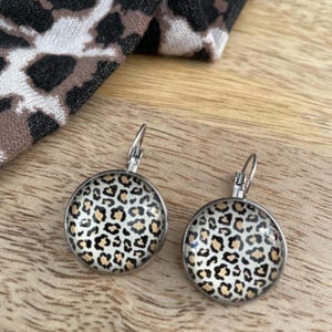Puede incluir: Un par de pendientes de aro de plata con un dije redondo de vidrio transparente con un diseño de estampado de leopardo marrón y negro.