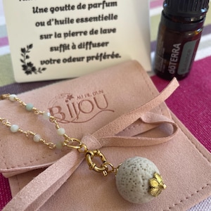 Peut inclure: Un collier doré avec un pendentif en pierre poreuse gris clair et de petites perles vert clair. Le collier est présenté avec une pochette rose et un flacon d'huile essentielle. Une carte avec du texte français est également visible.