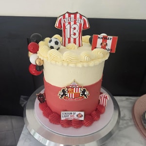 Peut inclure: Un gâteau à étages avec un glaçage crème et rouge, décoré de l'emblème du Sunderland A.F.C., d'un ballon de football, d'une figurine de maillot TYLER-JAY 13, d'un drapeau HAWAY THE LADS et d'une plaque STADIUM OF LIGHT SR5 SUNDERLAND A.F.C.
